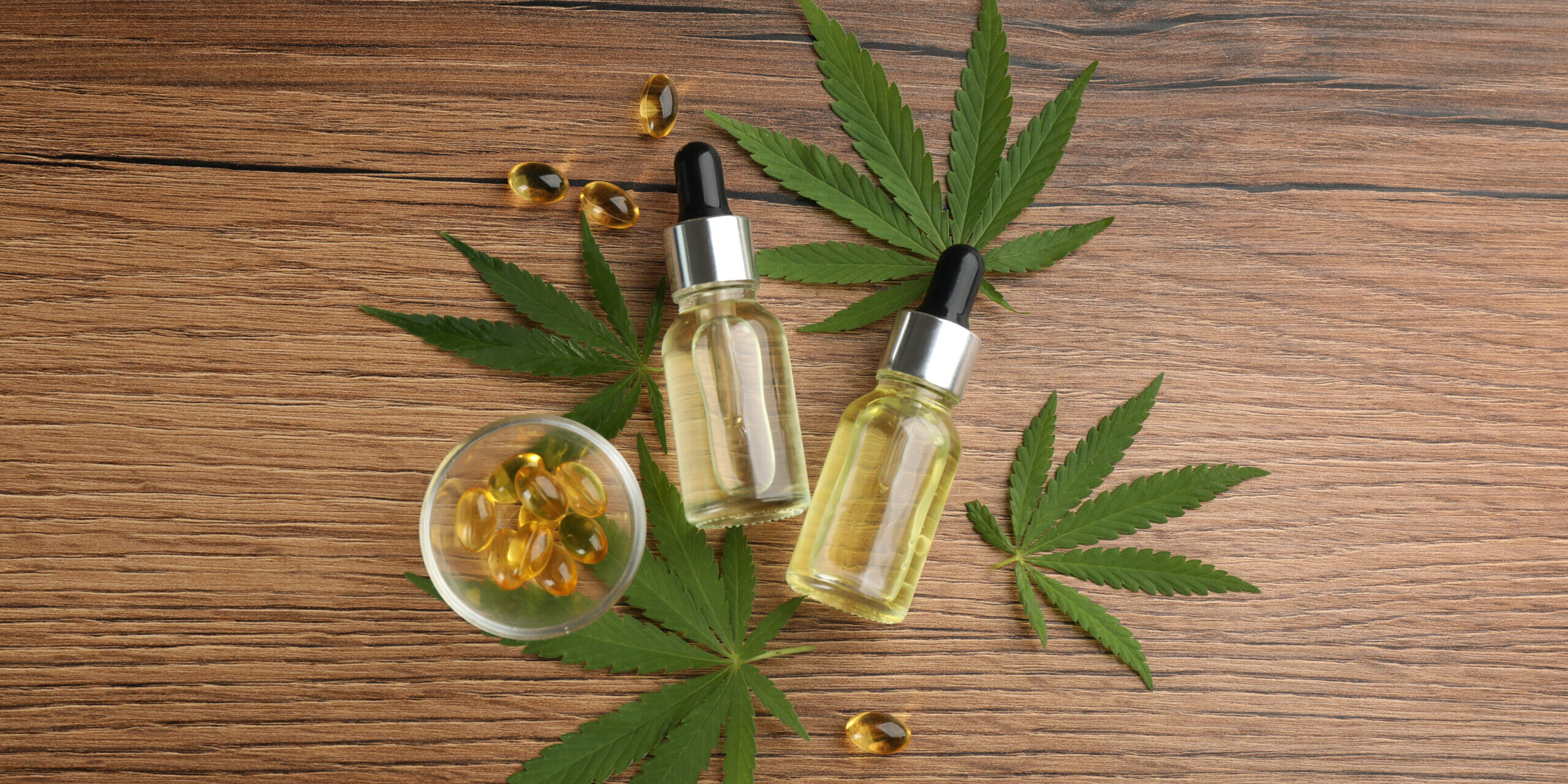 Two glass dropper bottles of cannabis tincture and a small bowl of softgel capsules arranged with cannabis leaves on a wooden surface, representing options for sleep aids.